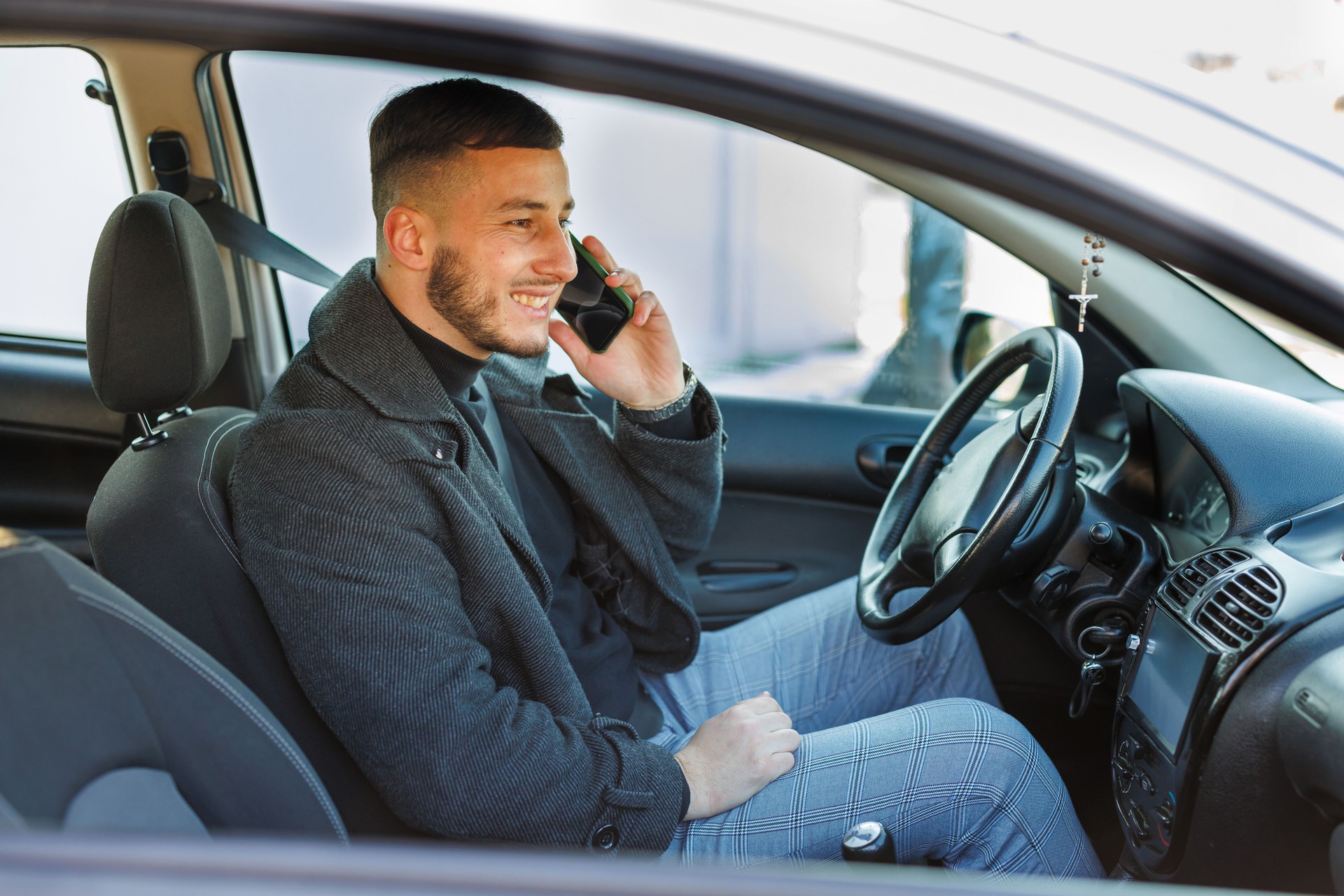 A young fashionable man is sitting in a car and talking using a phone.
