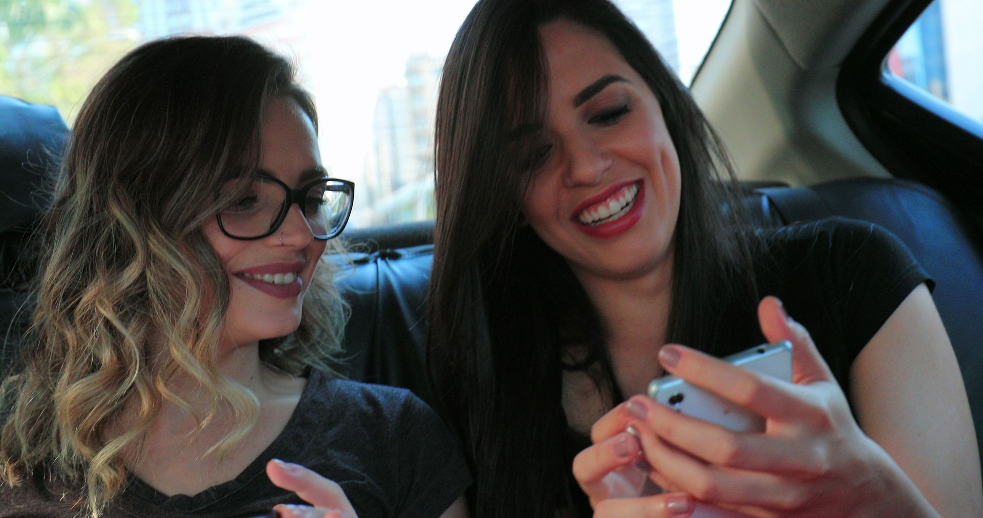 Candid girlfriends in the back seat of a car sharing cellphone screen showing content. Friends together riding taxi cab holding smartphone